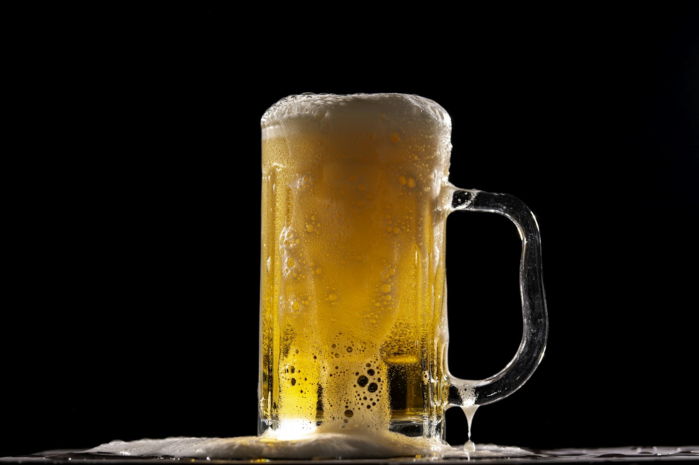 A foamy gold beer mug overflowing on a black backdrop